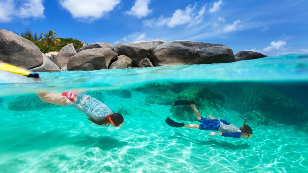 Two people snorkeling in clear, shallow water near large rocks under a blue sky with scattered clouds—a perfect scene for a family trip to Costa Rica.